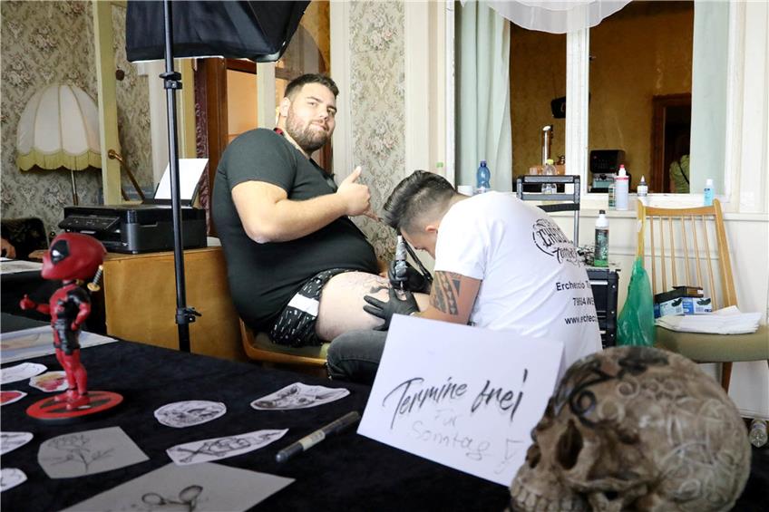 Ein verzierter Totenschädel auf dem Tisch. Im Hintergrund wird ein Wunsch-Tattoo auf den Oberschenkel fabriziert.